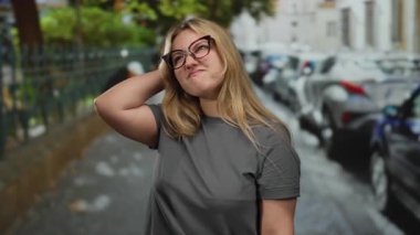 Gri tişörtlü ve gözlüklü bir kadın park etmiş arabaları ve arka planda yeşilliği olan bir şehir caddesinde dikiliyor düşünceli duygularını ifade ediyor..