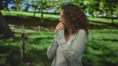 Elleri ağıza yapışan genç İspanyol kadın ve endişeli ifade ağaçların ve ahşap çitlerin yakınındaki bir orman parkında açık yeşil gömlek ve beyaz tank giyen endişeli yalnızlık.