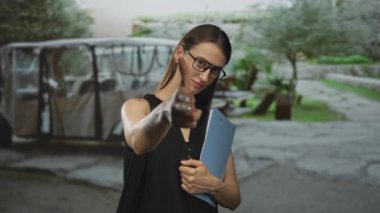 Siyah elbiseli kadın elinde mavi pano ile parketmiş karavan ve ağaçların yakınındaki caddede ileriyi işaret ediyor..