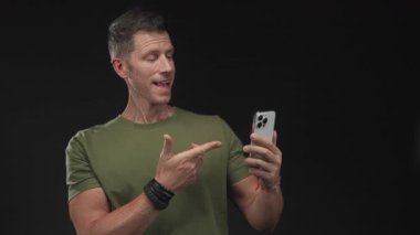 Adam siyah stüdyoda gülümserken ve telefonu tutarken akıllı telefonu işaret eder; güven bağlantısı.
