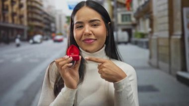 Kırmızı kutuda nişan yüzüğüyle dışarıda gülümseyen kadın, şehirde heyecan gösteriyor..