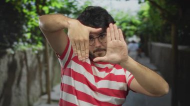 Sakallı ve gözlüklü genç bir adam sokakta elleri dışarıda, çizgili gömlek giyiyor, şehir ortamında odak ve yaratıcılık taşıyor.