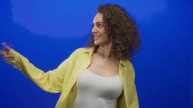 Mavi stüdyoda gülümseyen ve işaret ederken telefon eden, beyaz atlet üzerine sarı gömlek giyen bir kadın, mutluluk daveti.