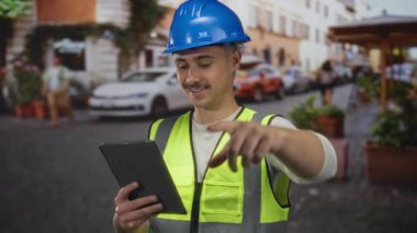 Şehir sokaklarında inşaat planlaması için tablet kullanan yansıtıcı yelek ve kasklı genç adam..