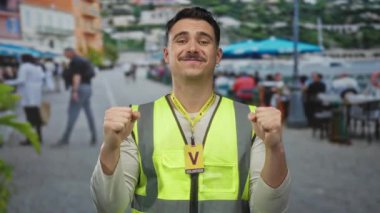 Young hispanic man in reflective vest volunteering on restaurant terrace with outdoor seating in vibrant area, expressing enthusiasm and positivity, surrounded by people enjoying coffee.