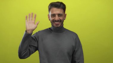 Gri balıkçı yaka kazak giyen İspanyol genç adam yeşil stüdyoda neşeli bir gülümsemeyle el sallıyor..