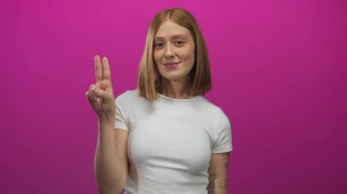 Kadın gülümsüyor barış işareti yapıyor. İki eli de açık pembe arka planda. Mutluluğu taşıyor, gençliği sarı saçlı, beyaz gömlekli, neşeli görünümlü..