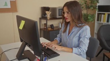 Gözlüklü ve mavi çizgili gömlek giyen bir kadın parlak ofis ortamında bilgisayarın çalışma yerinde klavyeye yazı yazıyor..