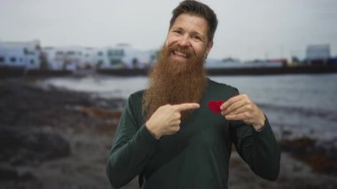 Adam arkasında kayalık bir sahil sahnesinde geniş bir gülümseme ve kırmızı bir kalp kesme tutarak başparmak verir; aşk.