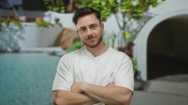 Sakallı genç adam arka planda yemyeşil ve modern mimarisi olan bir otelin havuzunun yanında gülümsüyor..