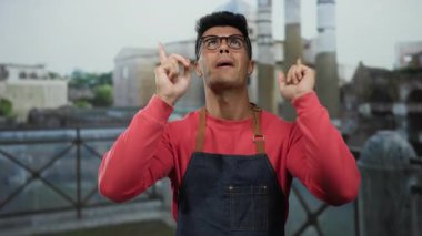 Gözlüklü ve kırmızı tişörtlü genç adam şehir caddesinde ellerini kaldırmış, bulanık mimari geçmişi ve kendine güveni ve neşesini sergiliyor..