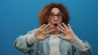 İspanyol, orta yaşlı, kıvırcık saçlı bir kadın kameraya, izole edilmiş mavi bir arkaplana, gözlüklü ve pozitif enerji gösteren günlük gömlekli bir kadına coşkuyla el sallıyor..
