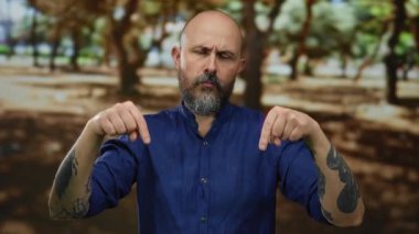 Mavi gömlekli kel bir adam parkta dikiliyor ve kollarında dövmeleri var..