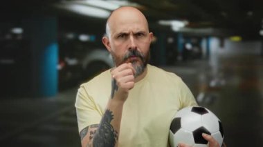 Kapalı otoparkta futbol topu tutan sakallı kel bir adam ciddi bir ifadeyle işaret ediyor ve kolunda dövmesi var..
