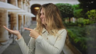 Sarışın, genç, gülümseyen bir avuç avuç içi açık havada bir ortam yaratırken kadın sütunlarda el hareketi yapıyor..