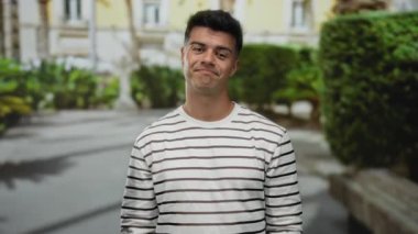 Çizgili tişörtlü İspanyol genç adam güneşli bir şehir caddesinde, şehir ortamında hoşnutsuzluğunu ya da anlaşmazlığını ifade ederek baş parmağını kaldırıyor..