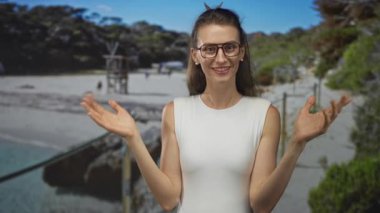 Gözlüklü ve çıplak omuzlu bir kadın, kumsalda neşeli bir jest içinde ellerini kaldırdı; neşeli bir doğallık.