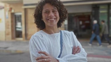 Woman standing with arms crossed on a city street, smiling broadly in natural daylight under clear sky; confidence.