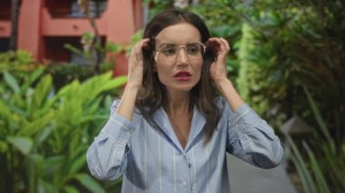 Kadın açık mavi gömlekle giriş yaparak yemyeşil yaprakların yanında dururken gözlük ve pürüzsüz saç ayarlar; merak.