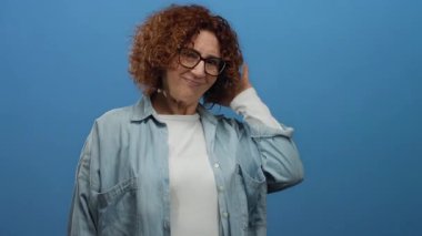 Gözlüklü bir kadın, mavi arka planda el ele gülümsüyor, sıradan bir kot gömlek ve kıvırcık esmer saçlar sergiliyor, rahat ve dostane bir atmosfer yaratıyor..