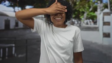 Beyaz t-shirt giyen kadın çizgi caddede gülümserken elleri ile gözlerini kapar; neşe dolu bir neşe.