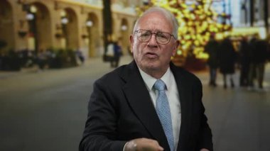 Takım elbiseli kıdemli bir adam hareketli bir açık hava caddesinde şenlikli şehir zeminine karşı canlı sohbetler yapıyor..