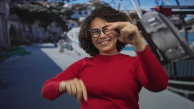 Gülümseyen kadın, parlak iskele rıhtımında, açık gökyüzü altında demirlemiş tekneler tarafından kamerayı işaret ediyor; güven güven güven bağı.