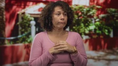 Pembe gömlekli kadın, parlak güneş ışığı altında bina girişinde avuçlarını birbirine bastırıyor; sakin yansıma meditasyonu.