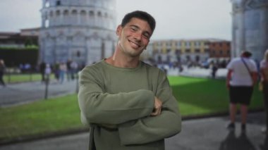 Genç adam gülümsüyor ve parmaklarını kameraya doğrultuyor caddedeki kuleye yaslanmadan önce; seyahat keşfi özgüveni.