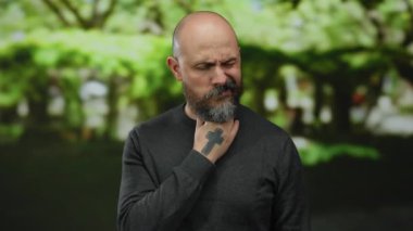 Dövmeli sakallı kel adam güneşli bir günde yemyeşil bir parkta boğazını tutmaktan rahatsız görünüyor..