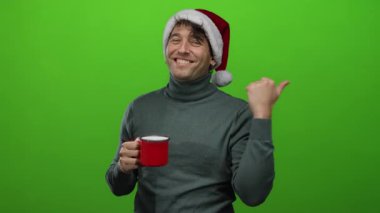 Noel şapkası takan İspanyol bir adam, neşeli tatil sahnesinde kırmızı bardağı canlı yeşil arka planda tutarken gülümsüyor ve başparmağıyla işaret ediyor..