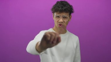 Gözlüklü ve beyaz gömlekli genç İspanyol adam mor bir stüdyoda kamerayı işaret ediyor; reddediyor..