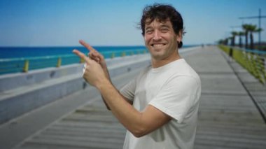 Beyaz gömlekli gülümseyen adam, arka planda açık mavi deniz ve gökyüzü olan güneşli sahil geçidini hevesle işaret ediyor..