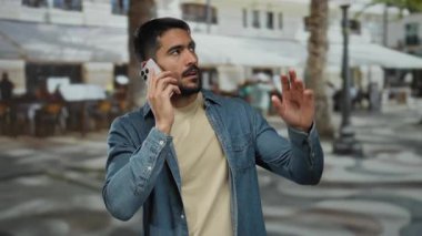 Şehir sokağında telefonla konuşan genç bir adam, günlük tişört ve bej tişört giyiyor, şehir geçmişi bulanık, açık hava endişelerini dile getiriyor..