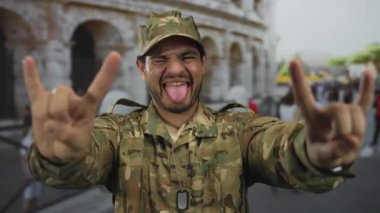 Kamuflaj üniformalı genç adam, dili dışarı çıkaran kaya boynuzlarını bina içinde hareket ettiriyor..