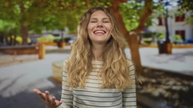 Kadın parkta konuşurken el kaldırırken, sarışın gülümseyen jestler açık hava kamera görüntülerini canlandırıyor..