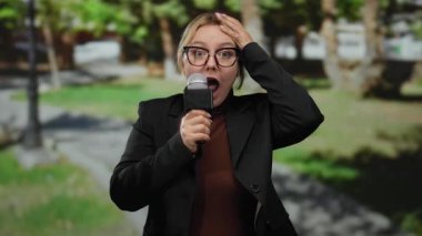 Açık hava parkında mikrofon tutan kadın muhabir arka planda yeşil ağaçlarla röportaj sırasında şaşkınlığını dile getiriyor..
