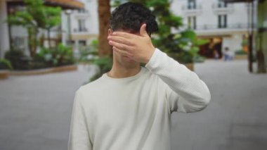 İnsan, şehir caddesinde gözleri kapamak için elini kullanıyor. Arkasında bulanık binalar ve yeşillikler var. Utanç..