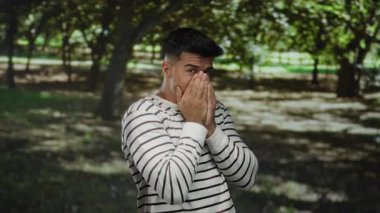 Çizgili tişörtlü genç adam endişeli görünüyor, güneşli bir parkta ağzı kapalı, gür ağaçlarla çevrili, sakin, açık havada derin düşüncelere dalmış bir ruh hali içinde..