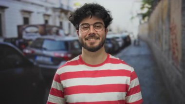 Sakallı ve gözlüklü genç adam arka planda park etmiş arabaları ve binaları olan bir sokakta gülümsüyor..