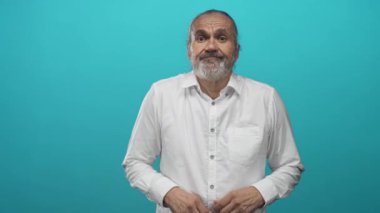 Uzun gri sakallı ve beyaz gömlekli uzun saçlı bir adam avuçlarını deniz mavisi atölyesinde silkeliyor; belirsizlik.