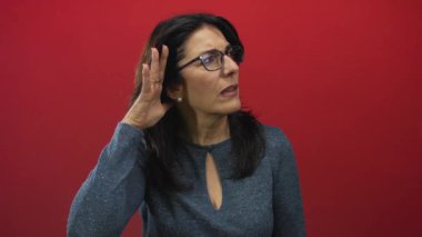 Orta yaşlı İspanyol kadın, gözlüklü, el pençe divan duran, stüdyoda dik dik bakan, merakla dinleyen..