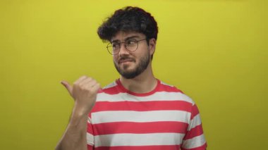 Parmakla işaret eden gözlüklü genç adam, parlak sarı arka planda çizgili gömlek giyiyor, rahat bir ifade ve oyuncu bir jest sergiliyor..