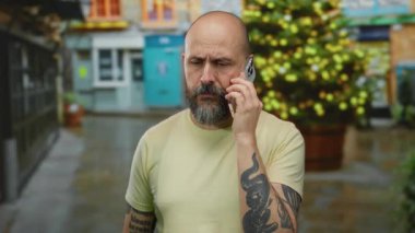 Sakallı bir adam şehir caddesinde telefonda konuşuyor. Noel ağacı arkaplanı var..
