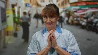 İspanyol kıdemli kadın hareketli bir şehir caddesinde sıcak bir şekilde gülümsüyor, açık şehir ortamında kendini adamış ve sıcaklığı somutlaştırıyor, insanlar ve binalarla çevrili..