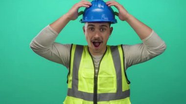 Yansıtıcı yelek ve başlık giyen İspanyol genç adam izole edilmiş yeşil bir arka plan duvarının önünde duruyor, şaşkınlığını dile getiriyor ve kafasını tutuyor..