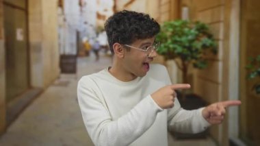 Genç İspanyol gözlüklü adam, taş binalarla dolu dar güneş ışığıyla aydınlanmış kaldırım taşı caddesinde parmağıyla işaret ediyor..