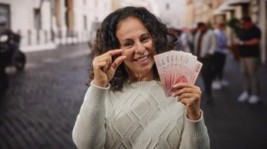 Bir sokak ortamında Tayvan banknotları tutan bir kadın, parmaklarıyla küçük bir jest yapıyor, insanların bulanık geçmişiyle şehir ortamında gülümsüyor..