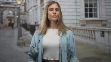 Genç sarışın bir kadın, gösterişli bir binayla sokakta elini kaldırmış ve ciddi bir ifadeyle işaret ediyor..