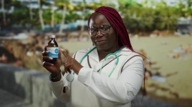 Deniz kenarındaki plajda elinde ilaç steteskobu tutan kadın gülümseyen Afro-Amerikan açık hava tıbbi, canlı, profesyonel arka plan.